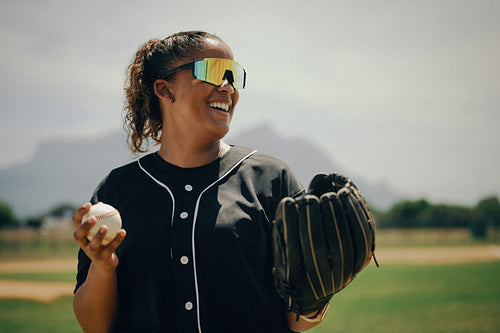 Cheerful baseball catcher in sunglasses stands on the field with glove and ball