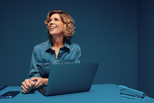 Mature businesswoman smiling and looking away while working on laptop and smartphone in monochromatic studio shot