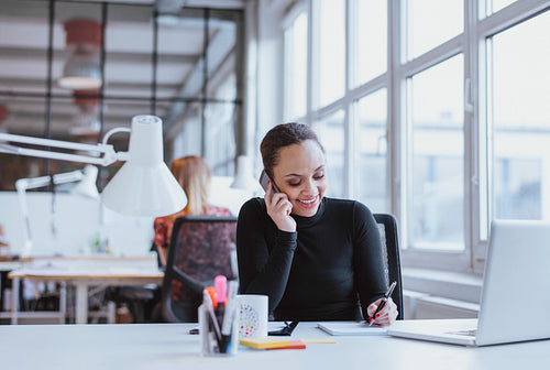 Happy young woman taking notes while talking on mobile phone