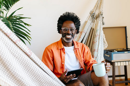 Relaxed black man sitting in a hammock with a cup of coffee and a touchpad