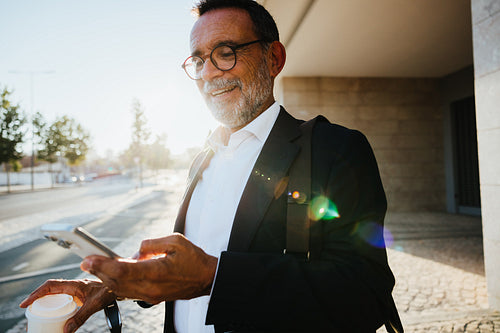 Senior businessman enjoying coffee while using smartphone outdoors in the morning