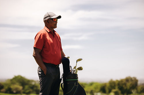 Male golfer wearing sunglasses watching over the golf course with a golf bag full of clubs