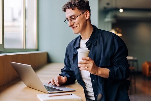 Young man working on laptop holding a coffee cup with a smile