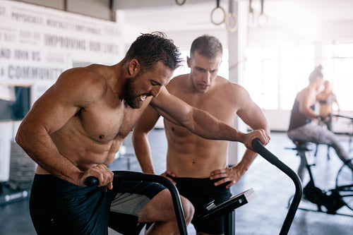 Muscular athlete doing intense workout on gym bike with coach