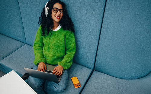 The woman relaxes on the sofa with a laptop, headphones, and smartphone at home