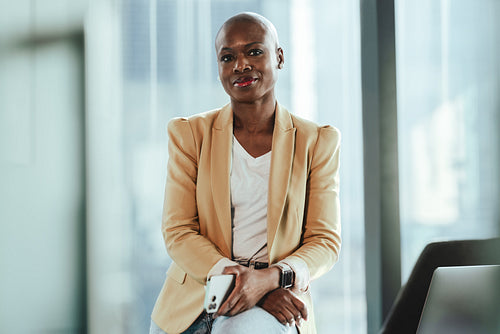 Confident mature businesswoman sitting in office, holding smartphone and looking at camera