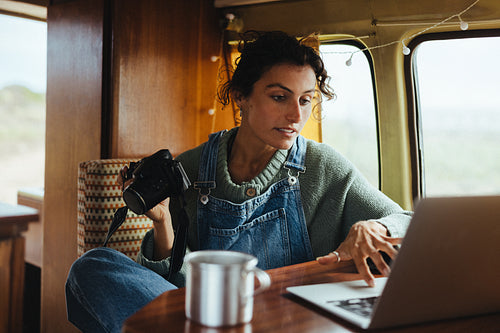 A young female photographer edits photos on a laptop inside a cozy camper, holding a camera