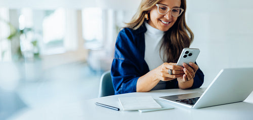 Woman using phone at desk with laptop