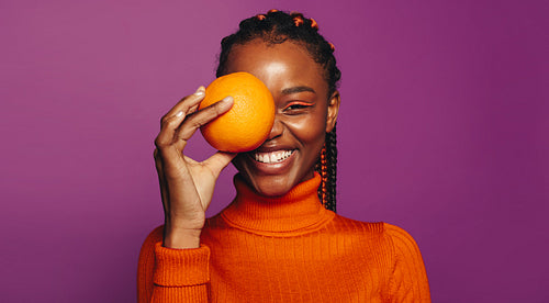 Happy african woman with colorful braids holding orange fruit on purple background