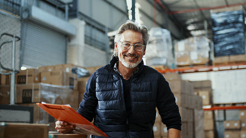 Mature and experienced warehouse manager smiles at the camera after reviewing inventory management records in a distribution center