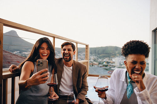 Friends enjoying wine and laughter while capturing fun moments on a balcony