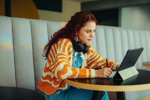 Focused female student studying with a tablet and headphones in a cozy, modern cafe setting