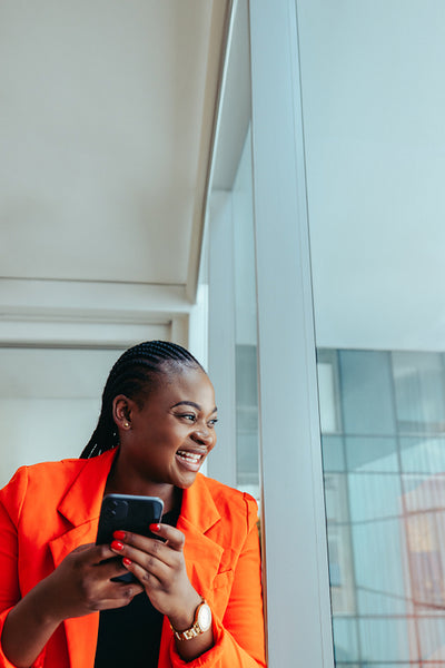 Smiling black woman in a bright orange blazer at the office in the city while using her smartphone