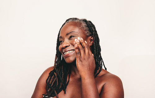 Cheerful black woman applying moisturizing cream on her face