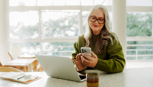 Senior woman uses smartphone beside laptop at home