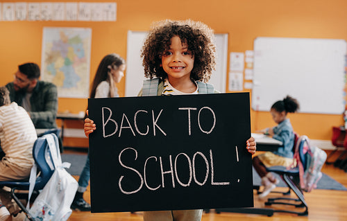 Proud pupil standing in a co-ed classroom on first day of elementary school, holding a back to school sign