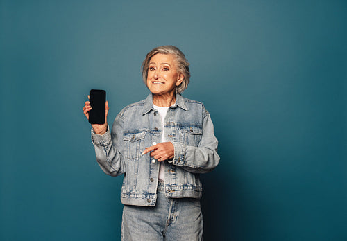 Senior woman recommending mobile app, holding up a smartphone in a studio