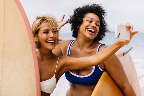 Happy female surfers taking a selfie together on the beach, celebrating their vacation in a fun surfing destination