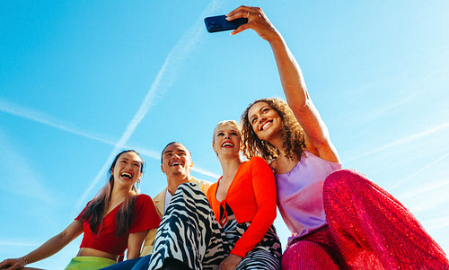 Young adult friends lean together as a smartphone captures them under a blue sky