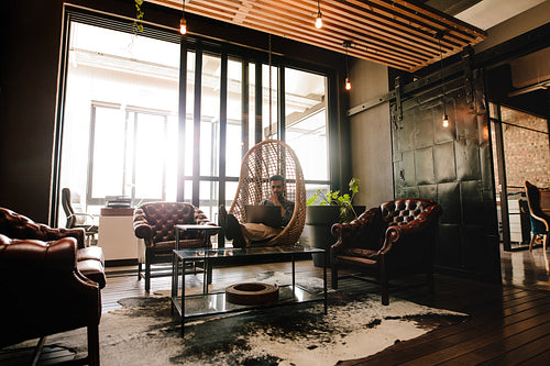 Man relaxing on hanging chair in office lounge with laptop