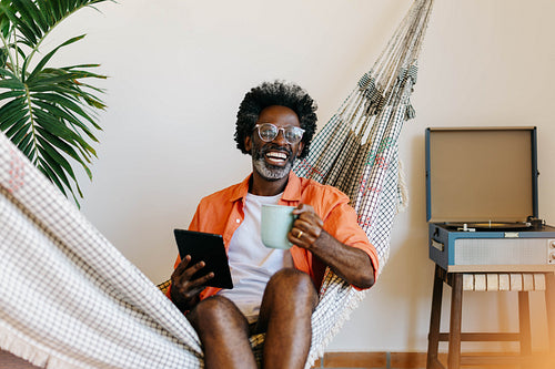 Happy black man relaxing in hammock with coffee and a tablet