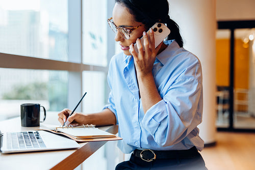 Focused woman in blue shirt on a call while taking notes at a desk