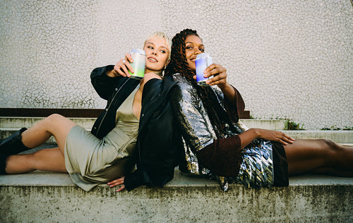 Women hold canned drinks while sitting back-to-back outdoors