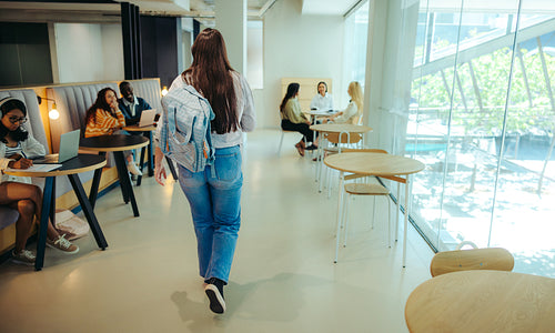 High school student walking through a modern library in the city, surrounded by peers studying with laptops and notebooks