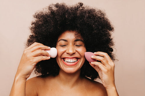 Happy young woman holding two make up sponges on her face