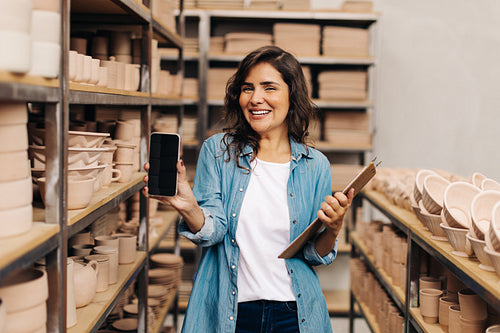 Happy ceramist showing a blank smartphone screen in her shop