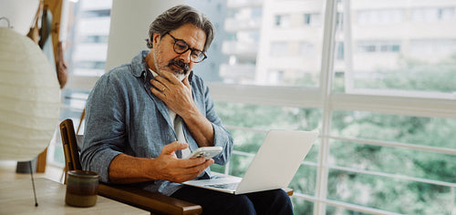Senior man uses smartphone with laptop at home