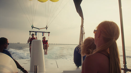 People parasailing over the ocean while friends watch from a boat during sunset