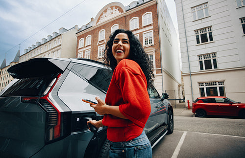 Smartphone in hand, a woman is charging her electric car on a city street