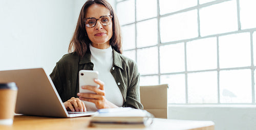 Woman using a mobile phone and a laptop to work in an office