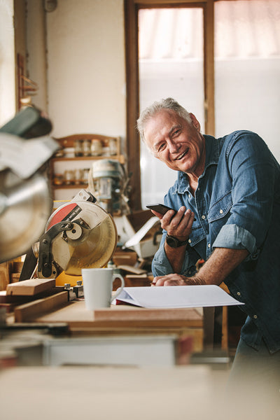 Happy senior male carpenter with phone in workshop