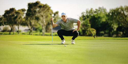 Focused male golfer crouching near a golf ball on the putting green on a picturesque golf course
