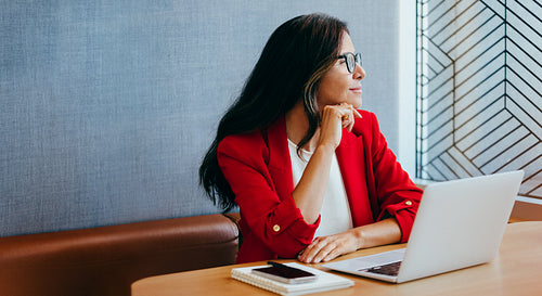 Businesswoman in bright red blazer working on laptop and looking out window