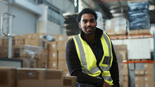Young warehouse employee smiling at the camera as he sits next to boxes of goods in a fulfilment center