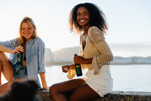 Joyful young friends enjoying beers by the waterfront at sunset