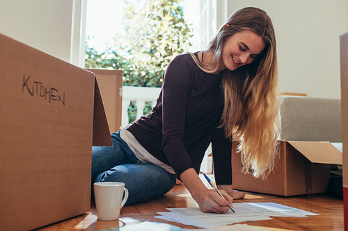 Smiling woman sitting with packing boxes on floor making a list