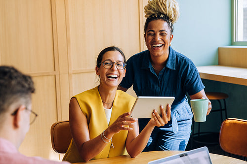 Two young professional women laughing while using a tablet in the workplace