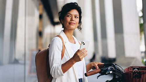 Thoughtful woman holding smartphone beside bike with basket on city street