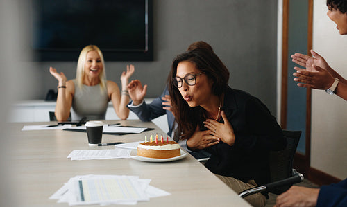 Business woman celebrating her birthday with her colleagues, blowing out candles on a cake in the office