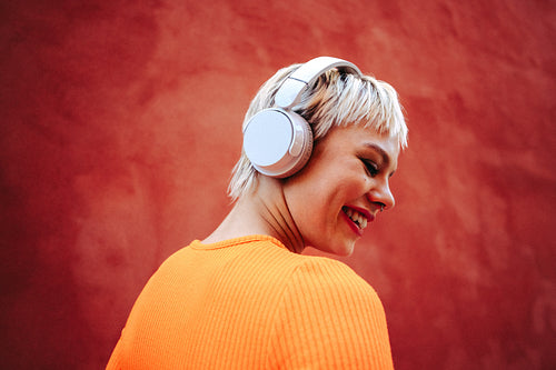 Woman smiles as she listens on headphones outdoors beside a red wall