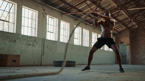 Muscular male athlete exercising with battle ropes in gym