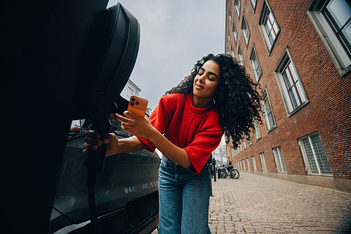 Woman charging electric car at public station in urban street while using smartphone