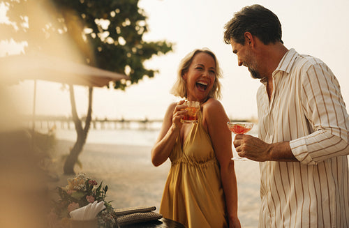 Couple enjoying cocktails by the beach at sunset, woman laughing