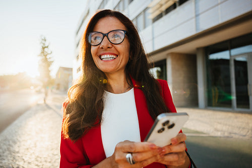 Smiling businesswoman holding a smartphone outdoors near an office building