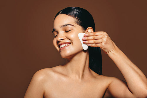Enjoying the gua sha magic: Happy young woman massaging her face with a gua sha stone