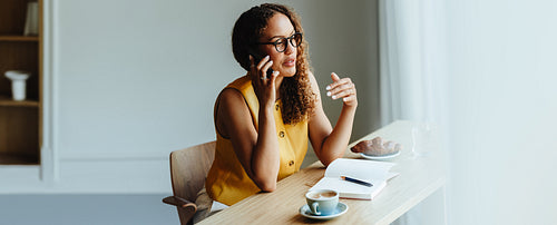 Woman sits at office table talking on the phone with coffee and notebook
